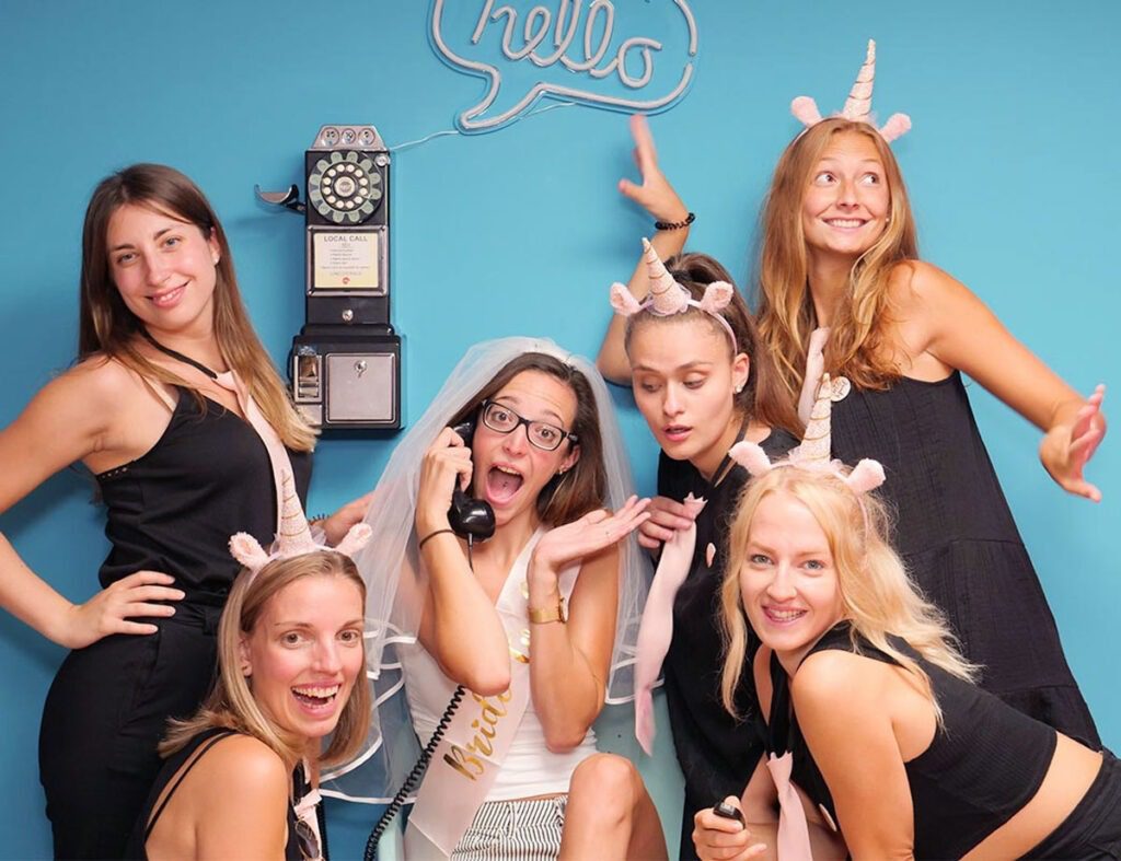 Fröhliche Braut mit ihrer Crew vor blauem Hintergrund und einem Münztelefon beim JGA Party Shooting